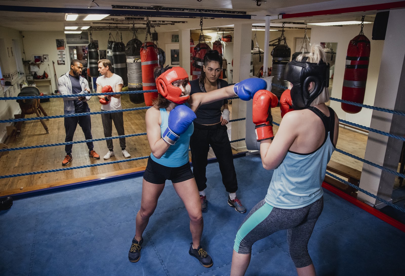 Two female amateur boxers mid-bout in a ring practising throwing punches and watched by female trainer. 