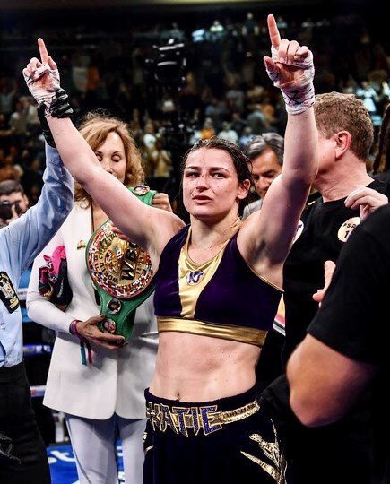 Katie Taylor pointing to the sky in victory with a lady holding her WBC championship belt.